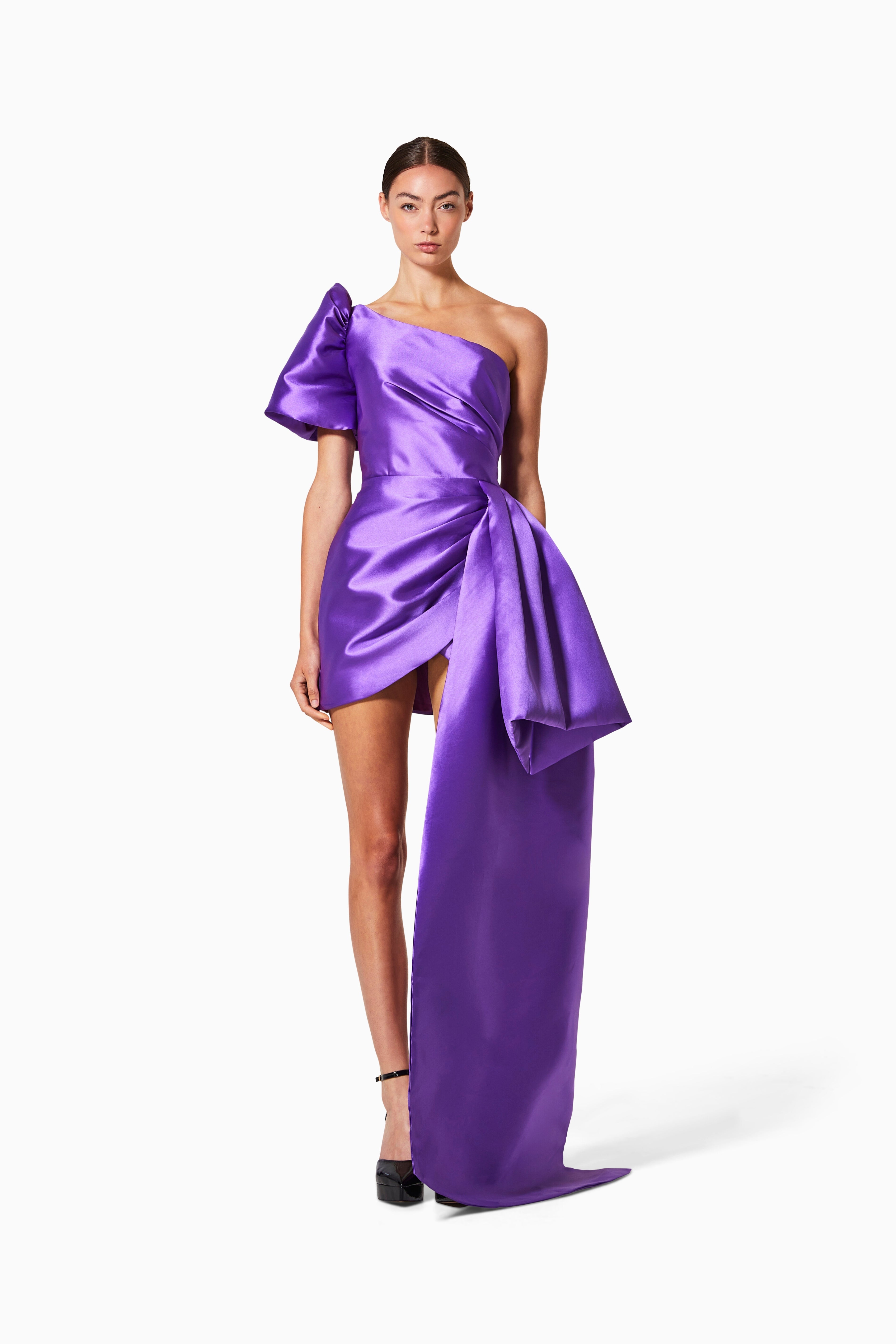 A model wearing a purple, lightweight pleated dress with side-sweep detail, standing against a plain background.