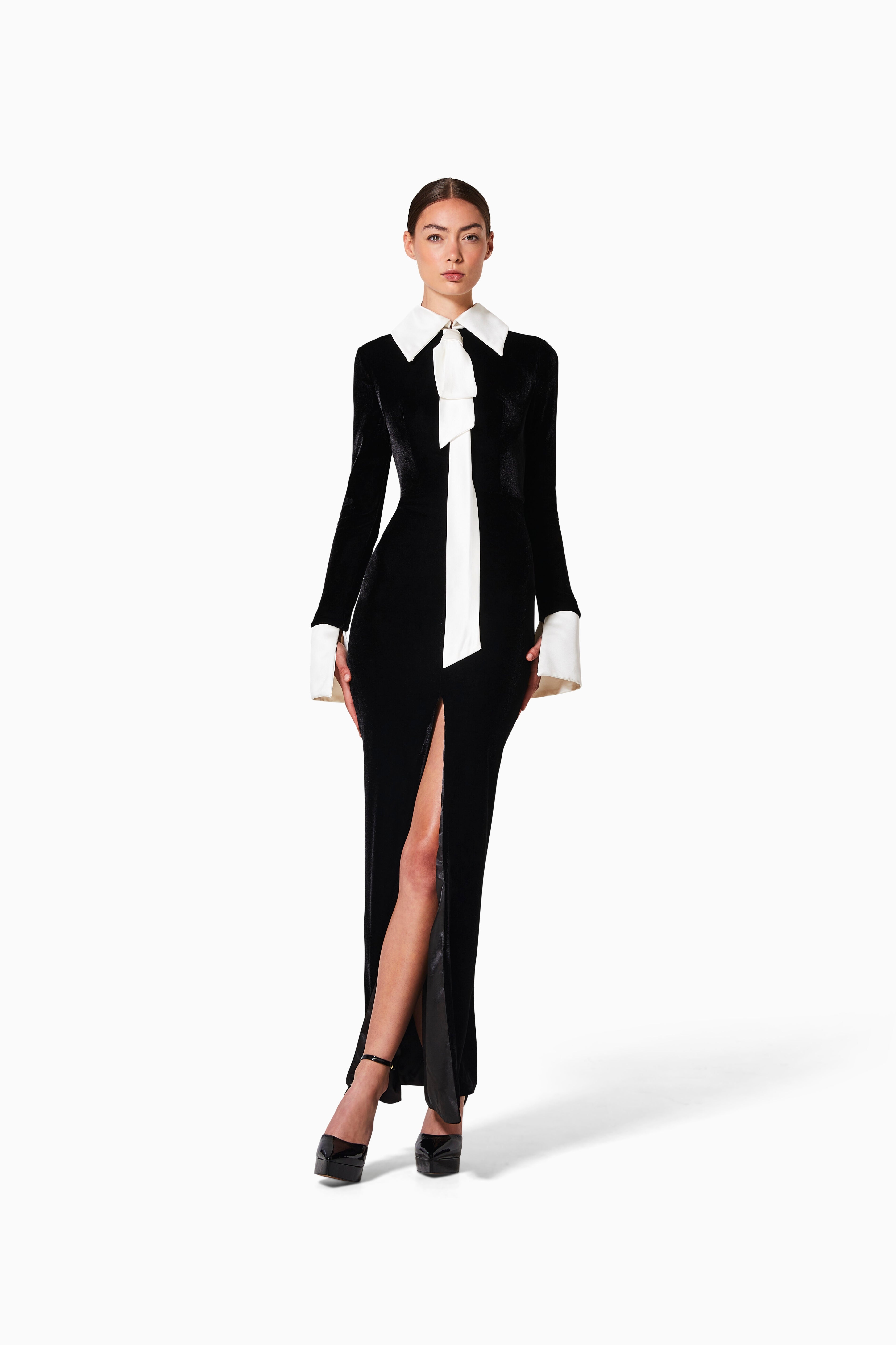 A model wearing a black velvet cocktail dress with silk neck and sleeves, featuring a high slit and tied detail.