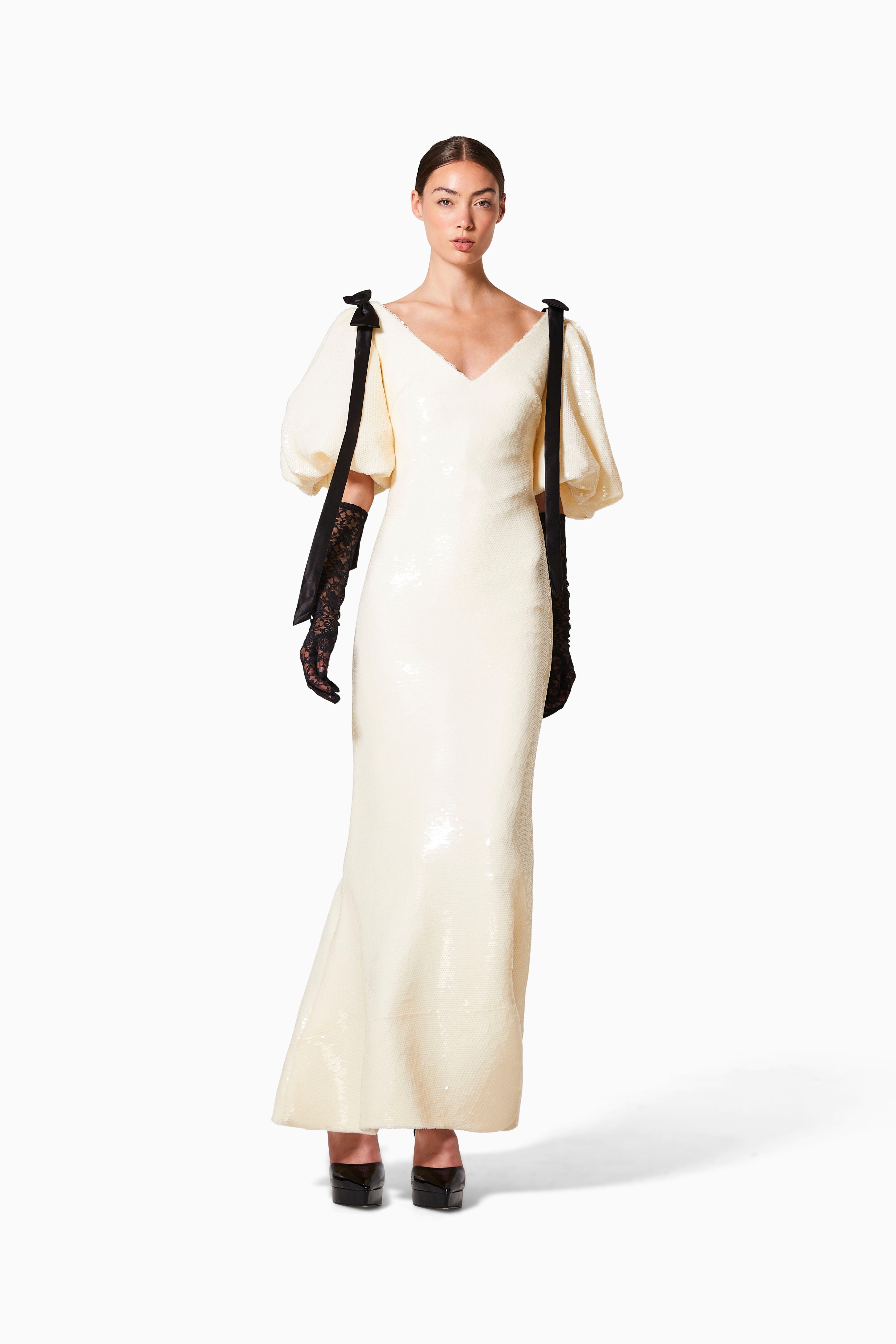 A woman standing wearing a cream sequin gown with puff sleeves and a bow detail on the shoulders.
