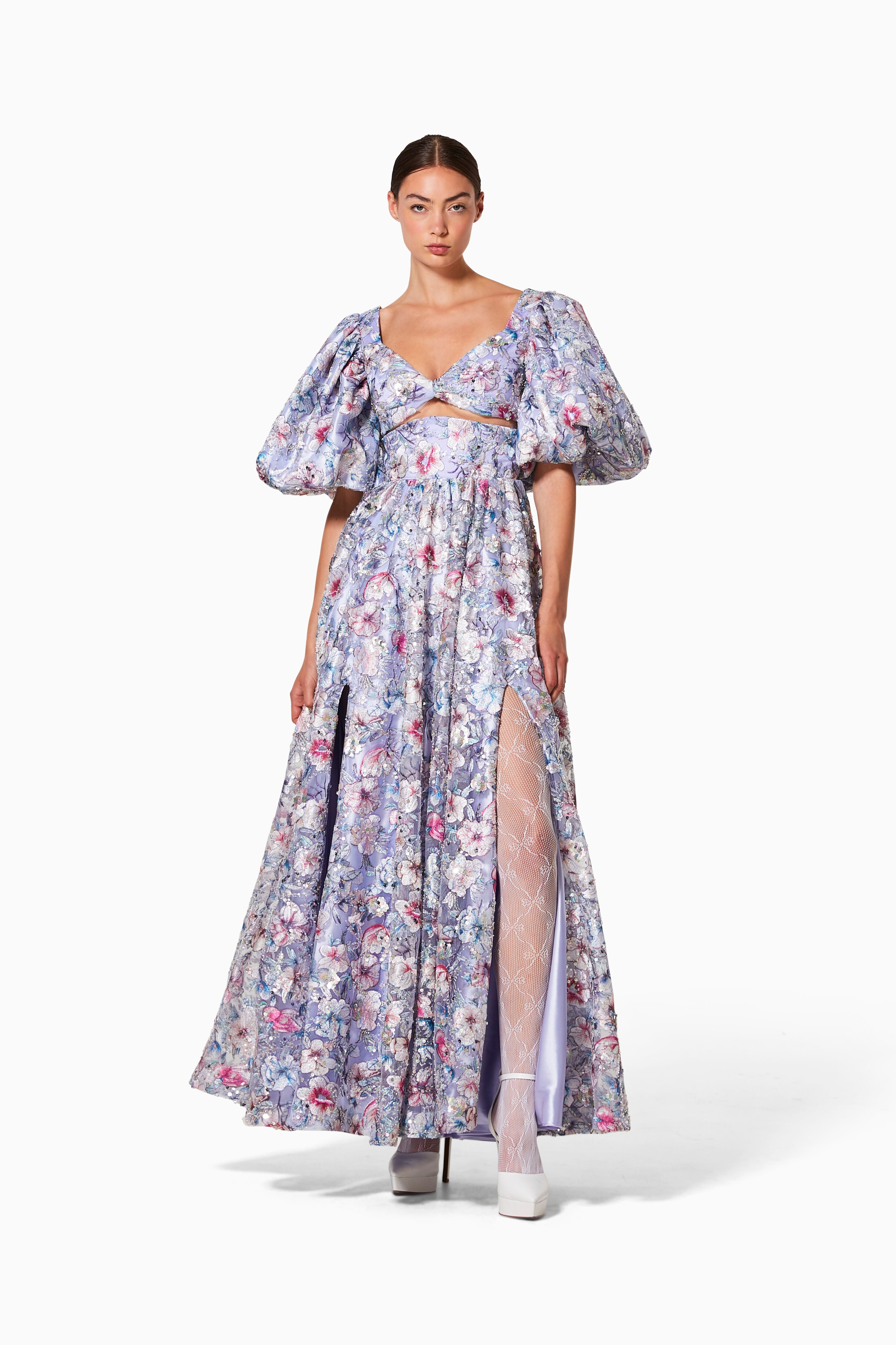 A woman standing wearing a flowing floral dress with front cutouts and juliet sleeves.